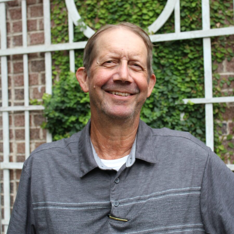 A man in a gray shirt smiles at the camera while standing in front of a brick wall with green vines and a white trellis.