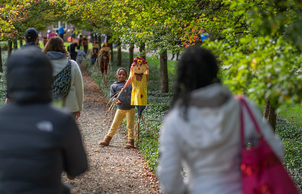 People walking along a tree-lined path with a child standing beside a scarecrow-like figure in the background.