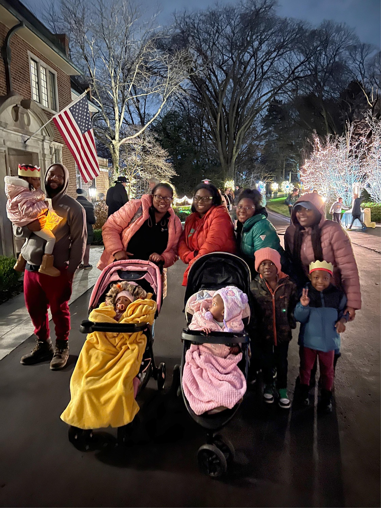 A group of people, including adults and children, stand outside at night next to strollers with toddlers. They are dressed warmly, and holiday lights decorate the background.