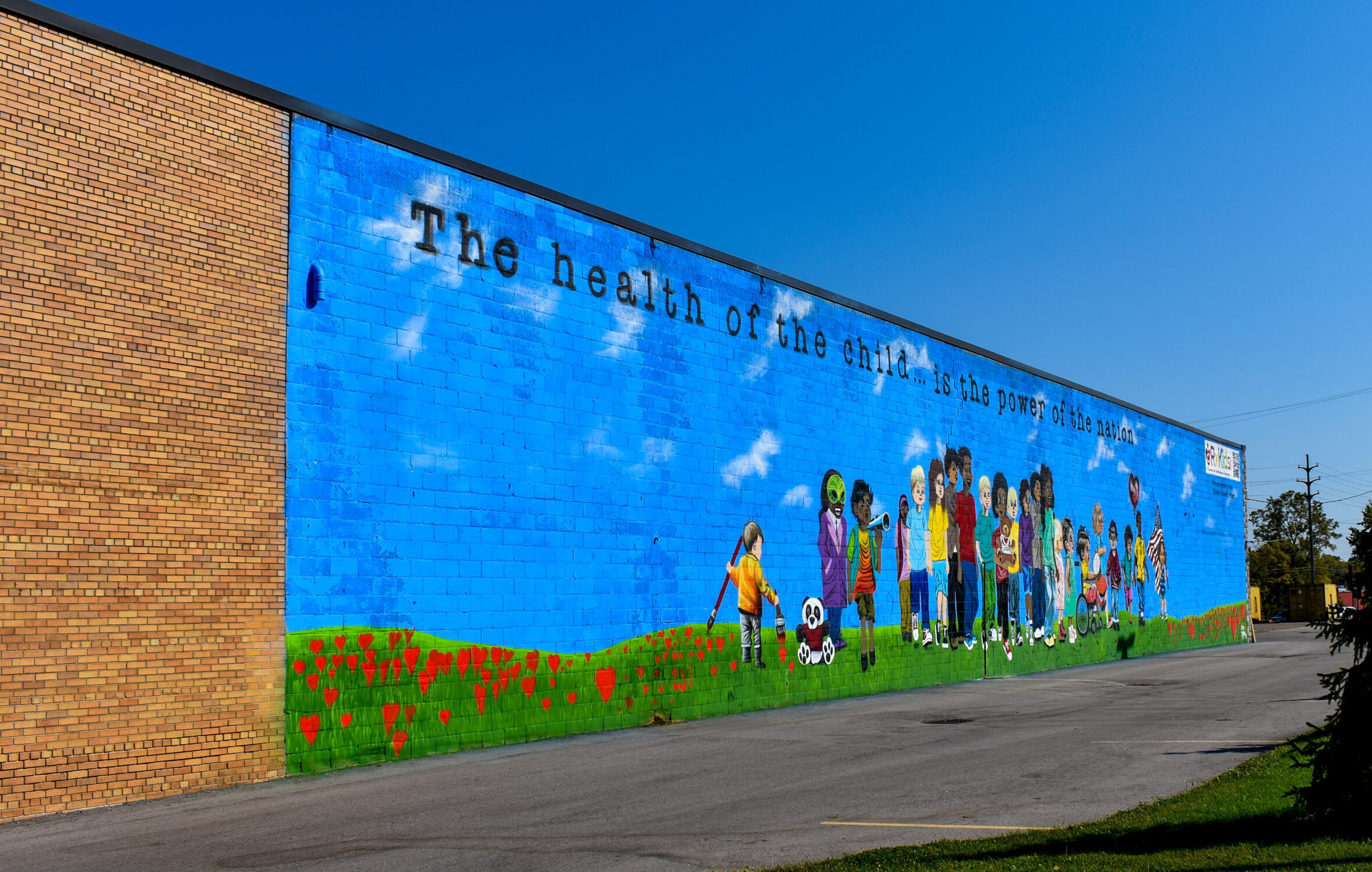 A colorful mural on a brick building, sponsored by the Ruth Mott Foundation, features a group of diverse children playing. Text above reads, 