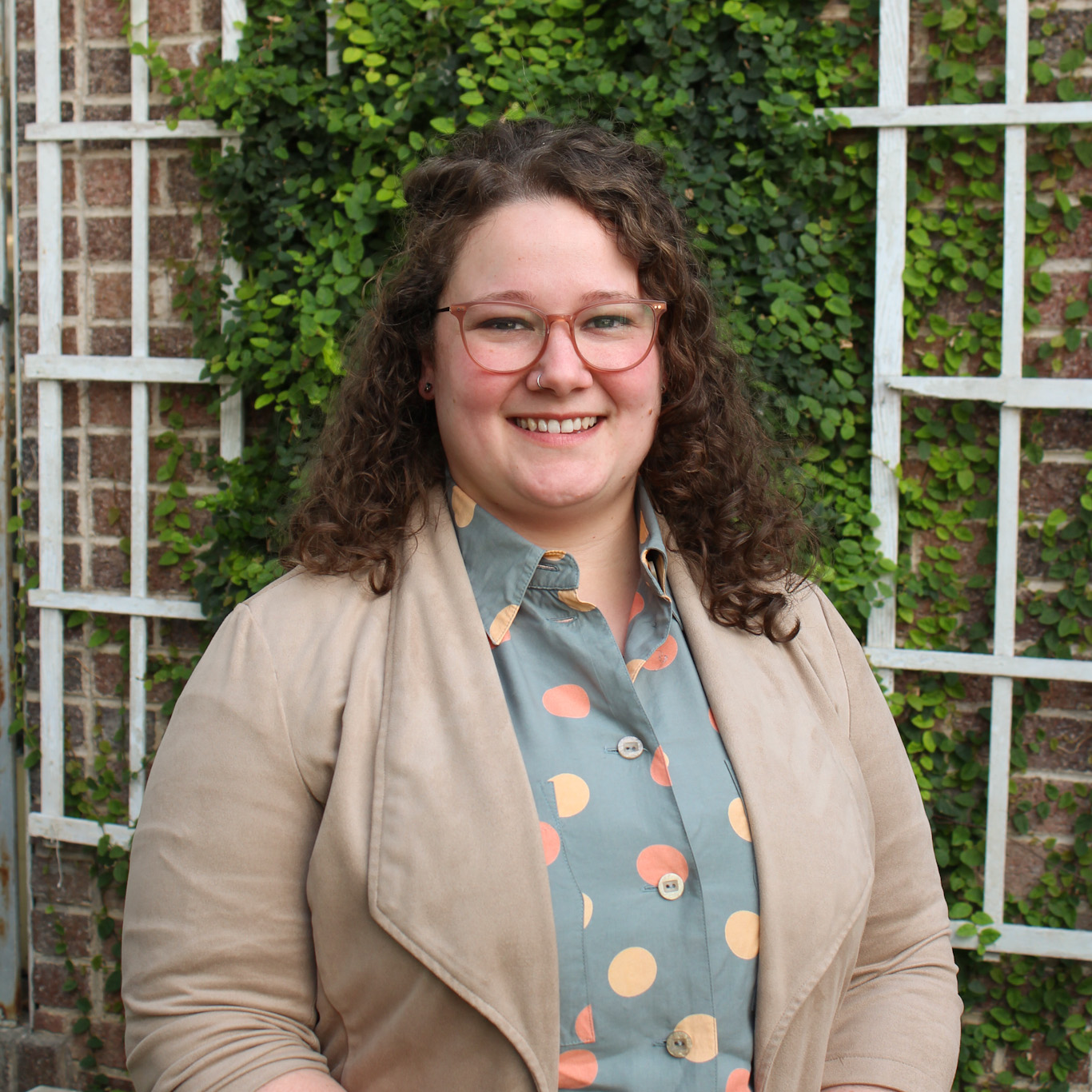 Person with curly hair and glasses smiling, wearing a polka dot shirt and light jacket, stands in front of a brick wall covered with green plants.