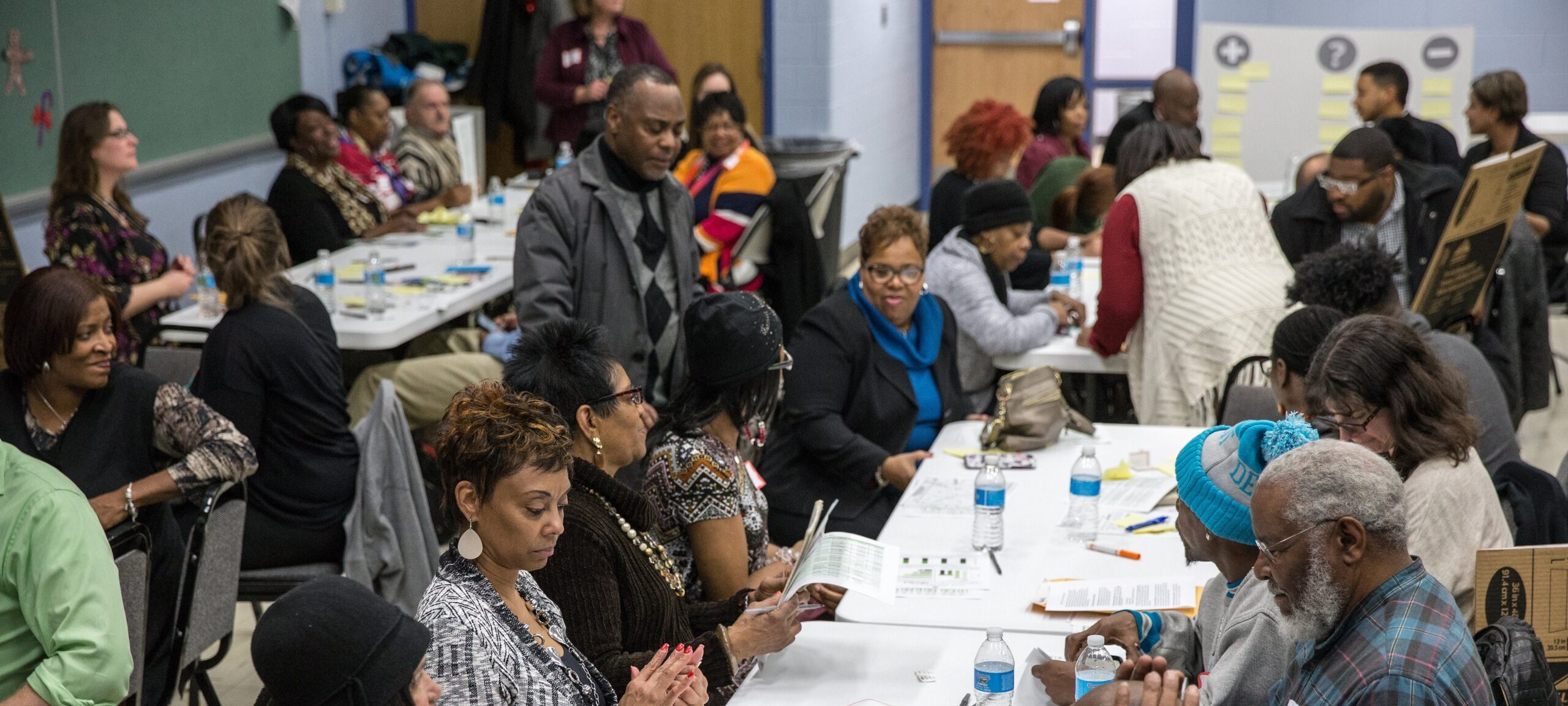 A diverse group of people seated at tables in a community meeting, engaging in discussions and reviewing materials.