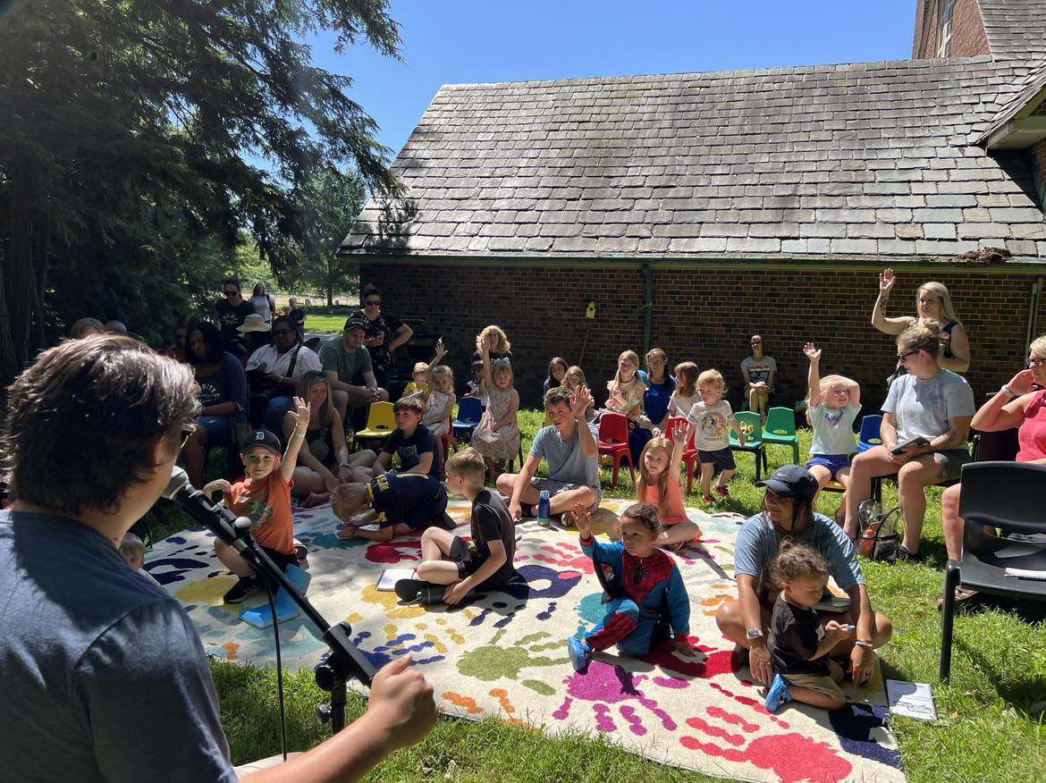 A person speaks into a microphone outside to a group of seated children and adults on colorful chairs and a patterned rug under sunny weather. Some children raise their hands.